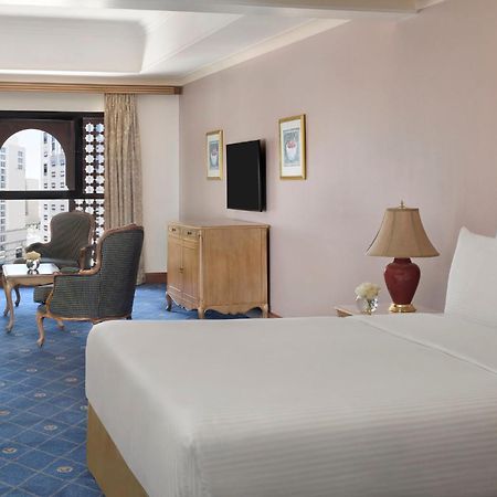 Intercontinental Dar Al Iman Madinah By Ihg Hotel Medina Kültér fotó The photo shows a well-furnished hotel room. It features a large bed with a white duvet, two bedside lamps, and decorative paintings on the walls. There is a comfortable seating area with two chairs and a small table. A wooden dresser is positioned a