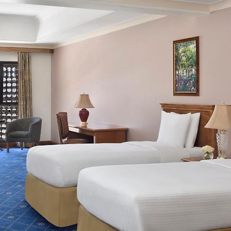Intercontinental Dar Al Iman Madinah By Ihg Hotel Medina Kültér fotó The photo shows a nicely designed hotel room featuring two twin beds with white linens and beige bases. In the background, there's a wooden desk and a lamp, along with a pair of comfortable armchairs positioned near a large window. The window showcas
