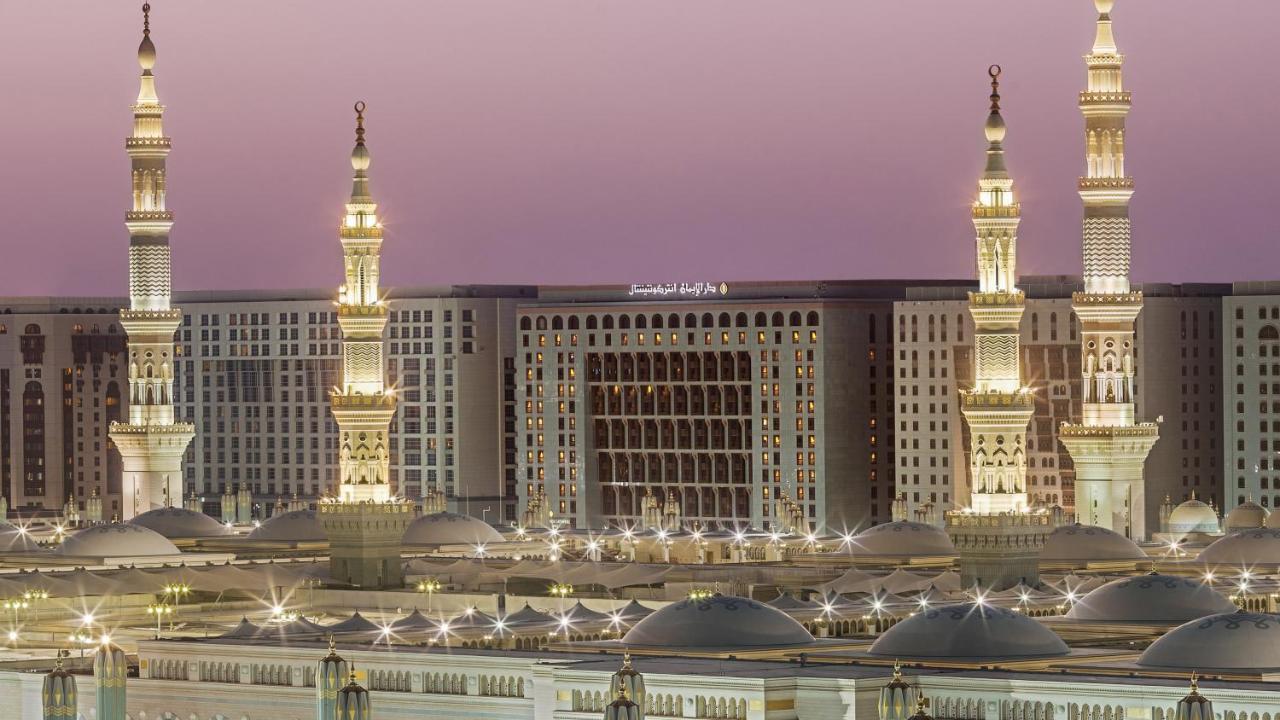 Intercontinental Dar Al Iman Madinah By Ihg Hotel Medina Kültér fotó The photo depicts an architectural scene, likely from a mosque complex, featuring prominent minarets that are illuminated. The setting appears to be during twilight, with a soft gradient in the sky. In the background, a large building is visible, pos
