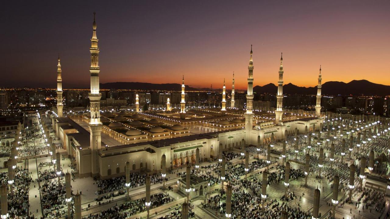 Intercontinental Dar Al Iman Madinah By Ihg Hotel Medina Kültér fotó The photo depicts an aerial view of a large mosque complex during sunset. The mosque features multiple tall minarets, which are intricately designed and illuminated, giving the scene a stunning appearance as the sky transitions from daylight to dusk.