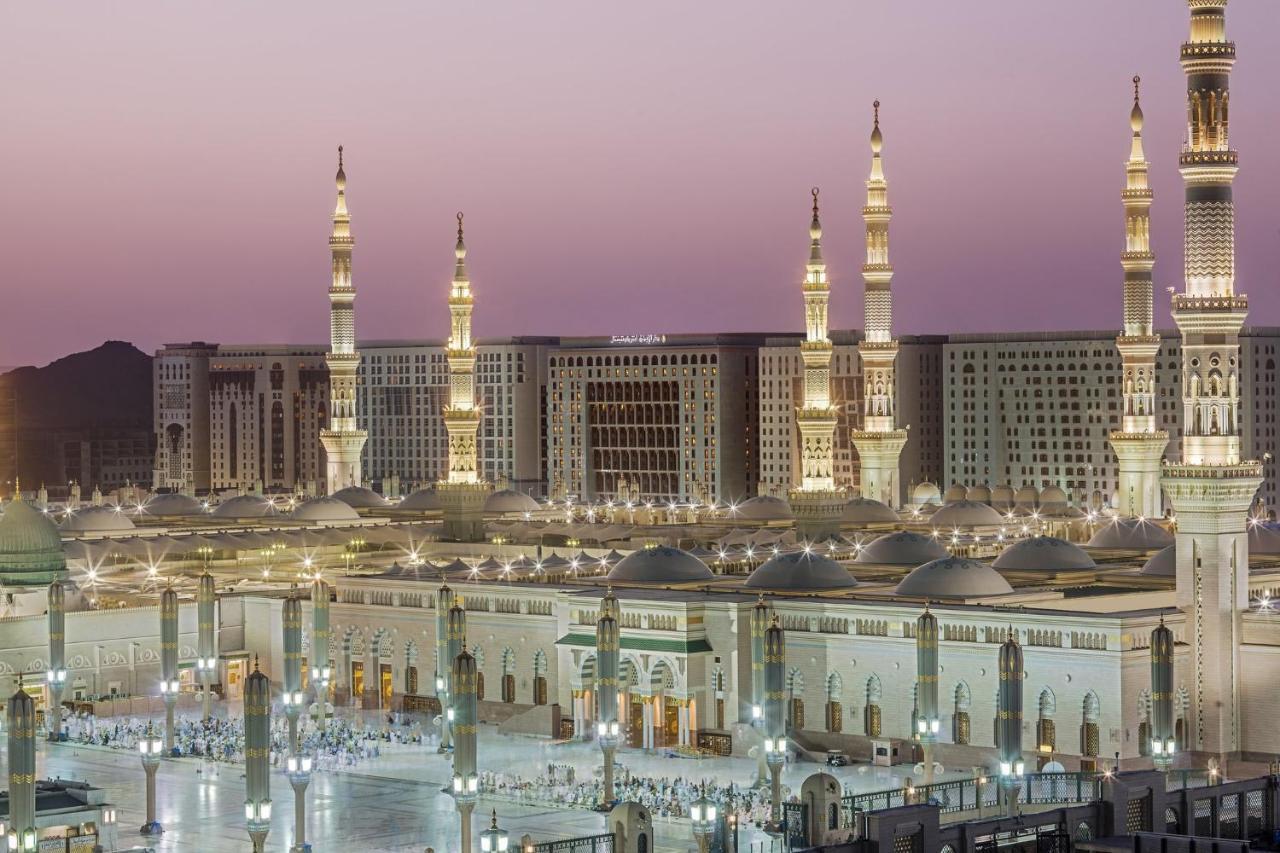 Intercontinental Dar Al Iman Madinah By Ihg Hotel Medina Kültér fotó The photo shows a view of an expansive mosque complex, featuring several tall minarets that are illuminated against a twilight sky. The structure appears grand and is likely adorned with intricate architectural details. There are people gathered in t