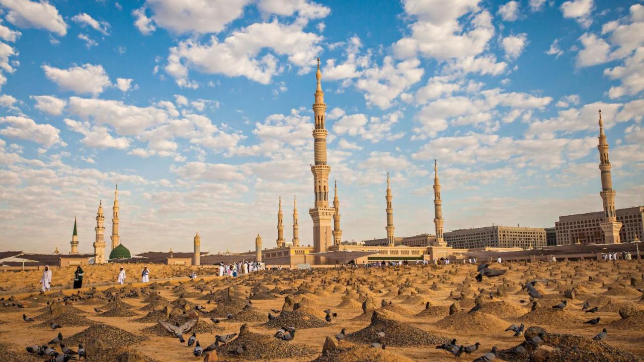 Intercontinental Dar Al Iman Madinah By Ihg Hotel Medina Kültér fotó The photo shows a landscape that features a large mosque with a tall minaret surrounded by smaller minarets. In the foreground, there are numerous grave markers arranged in a desert-like setting. The sky is bright with scattered clouds, and there are