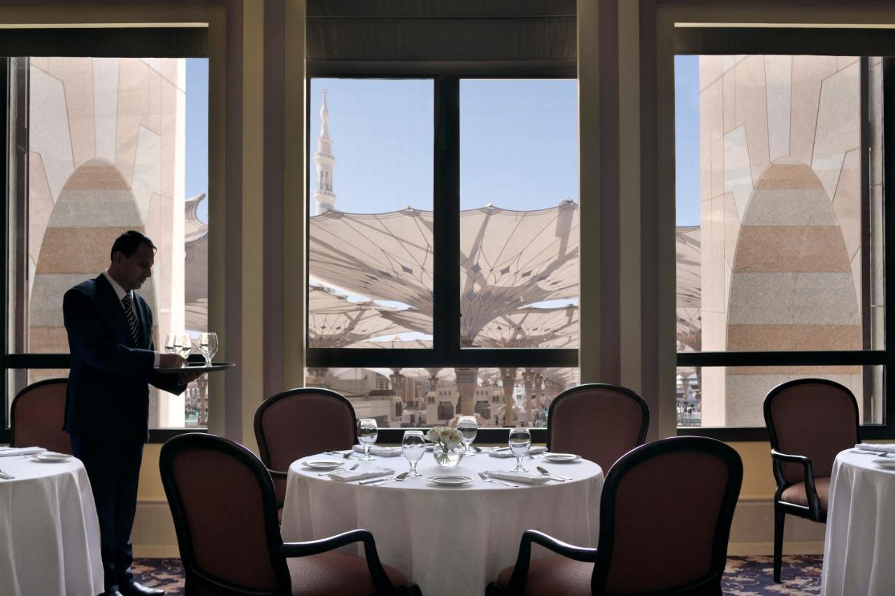 Intercontinental Dar Al Iman Madinah By Ihg Hotel Medina Kültér fotó The photo shows a dining area with elegant decor. A waiter is standing to the side, holding a tray. The tables are neatly set with white tablecloths and fine dining arrangements. Large windows provide a view of an outdoor area featuring architectural