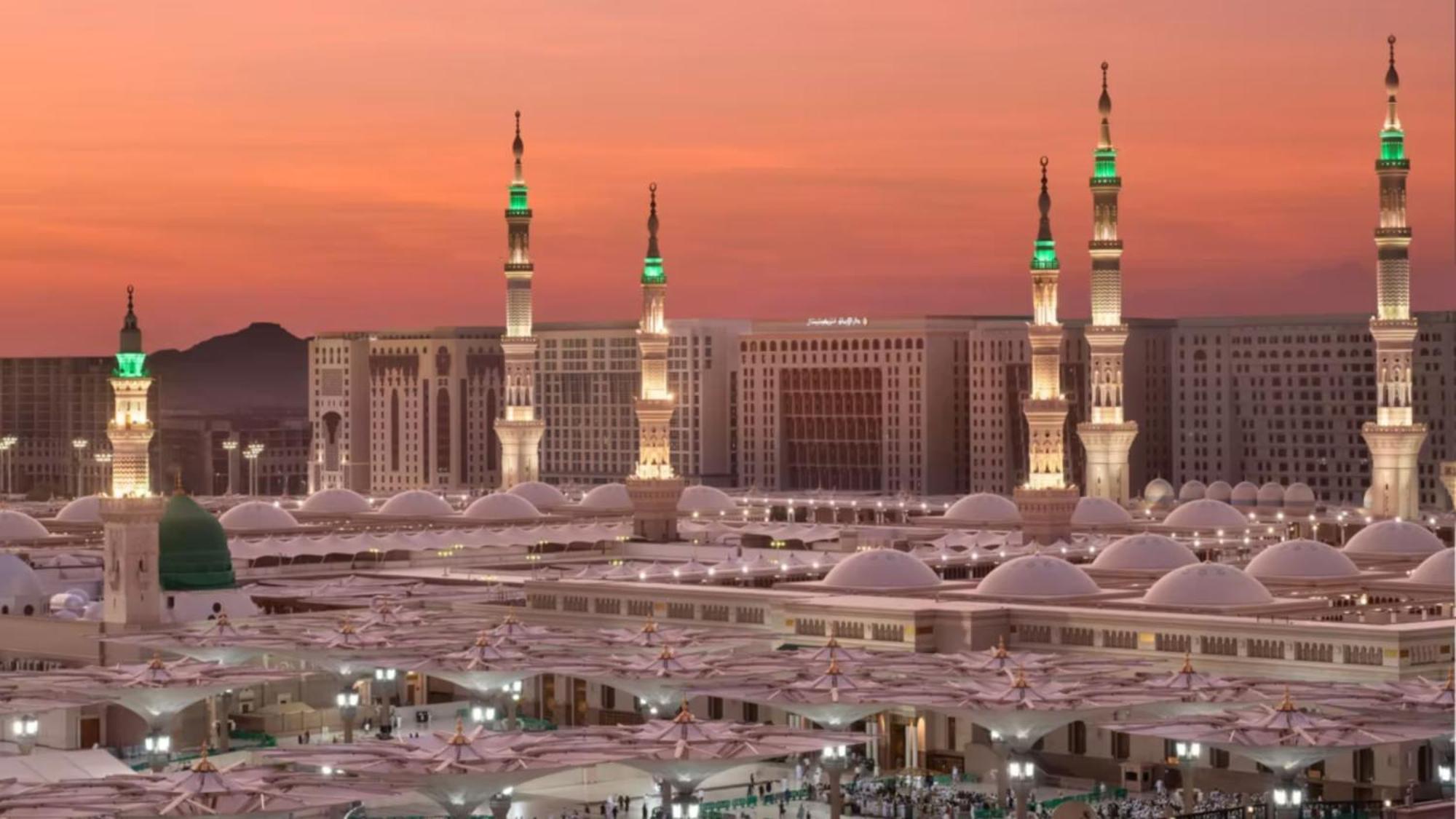 Intercontinental Dar Al Iman Madinah By Ihg Hotel Medina Kültér fotó The photo depicts a scenic view of a mosque complex, likely during sunset, with several minarets illuminated and showcasing green lights. The architecture features domed roofs, and the setting includes a backdrop of buildings, all bathed in a warm, o