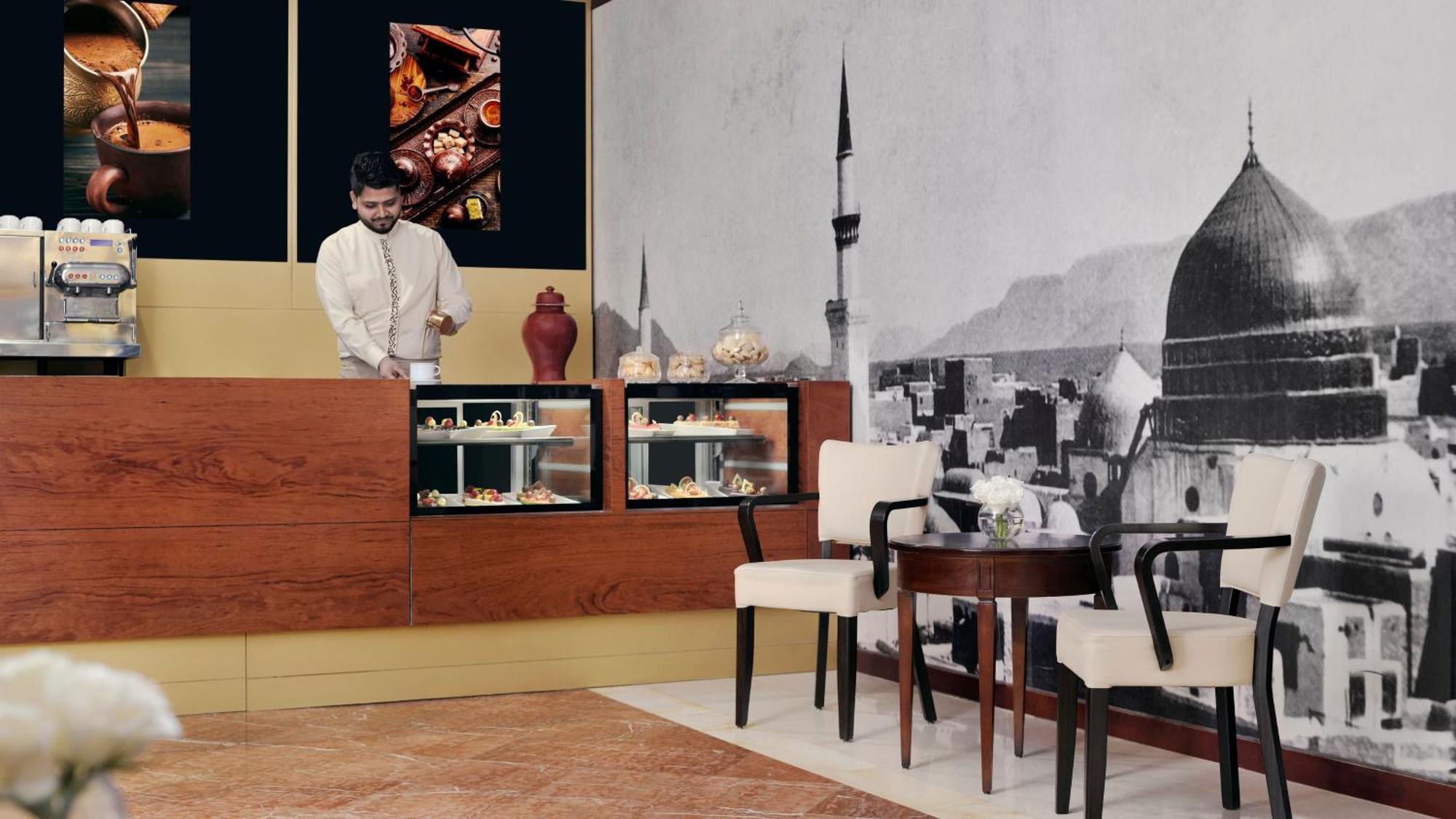Intercontinental Dar Al Iman Madinah By Ihg Hotel Medina Kültér fotó The photo shows an elegant cafe or dessert shop interior. A man in traditional attire is standing behind a wooden counter, where various pastries and sweets are displayed. The walls feature a large black-and-white mural of a cityscape, possibly depic