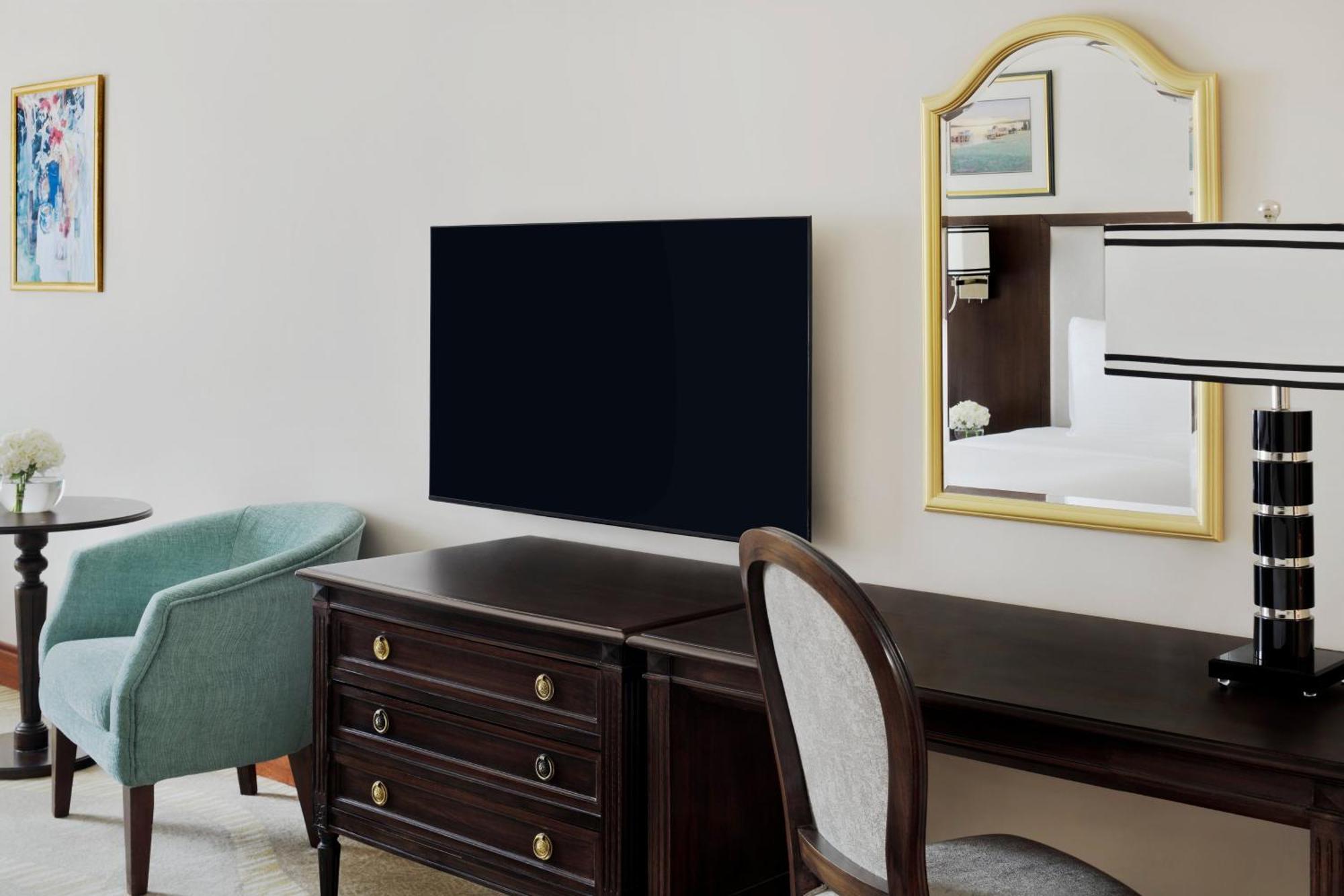 Intercontinental Dar Al Iman Madinah By Ihg Hotel Medina Kültér fotó The photo shows a hotel room interior featuring a mounted flat-screen television on the wall. Below the TV, there is a dark wooden dresser with drawers. To the right, there's a wooden desk and a chair with a light-colored seat. A small table with a l