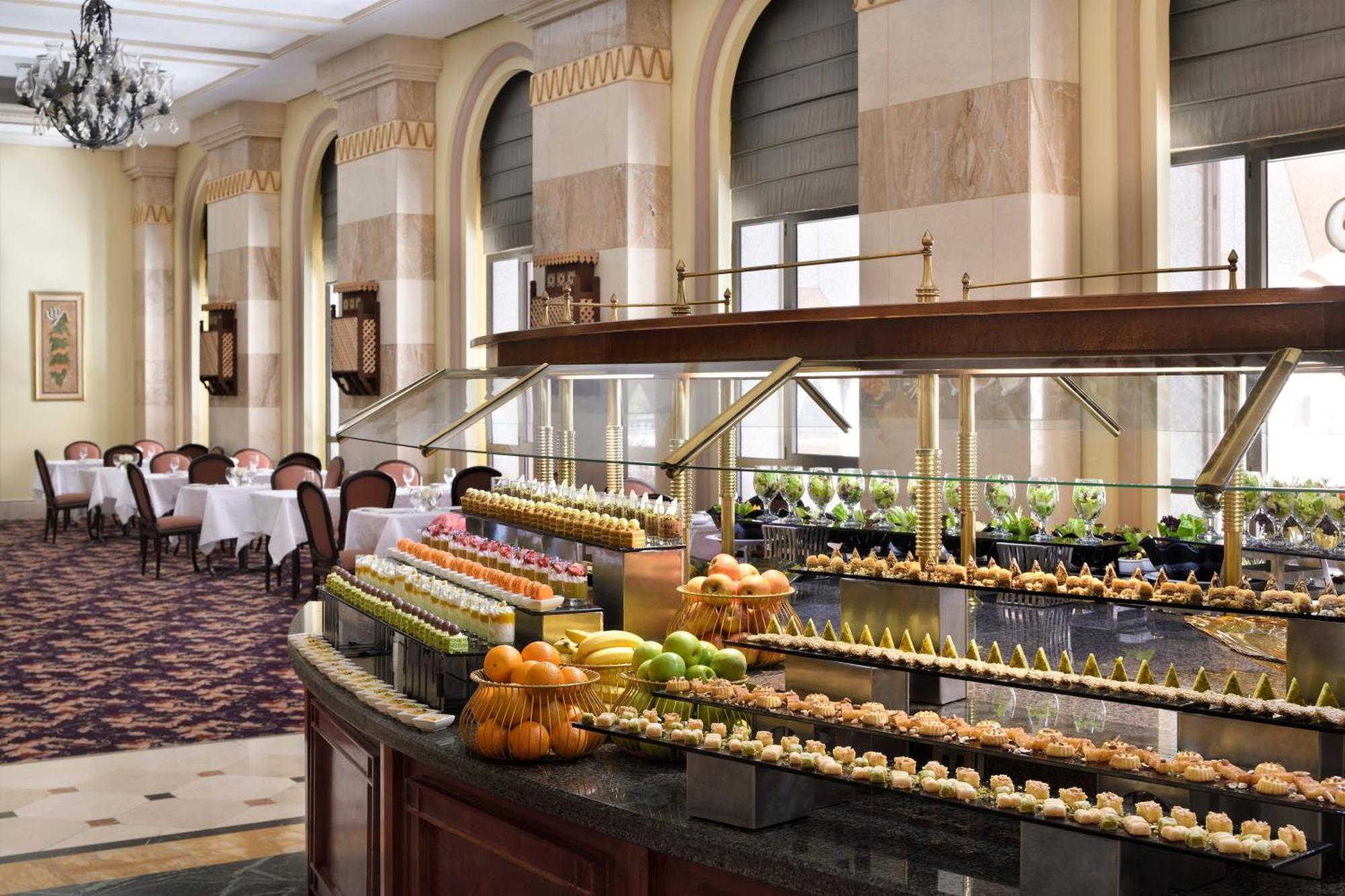 Intercontinental Dar Al Iman Madinah By Ihg Hotel Medina Kültér fotó The photo depicts an elegant dining area featuring a buffet-style display. In the foreground, a beautifully arranged buffet counter showcases an array of desserts, including miniature pastries and colorful fruit. The setting has a sophisticated ambia