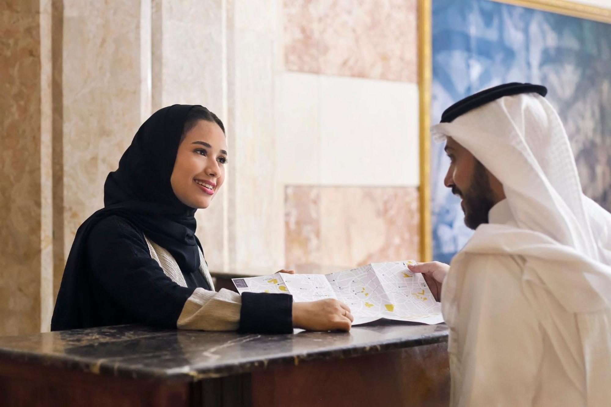 Intercontinental Dar Al Iman Madinah By Ihg Hotel Medina Kültér fotó The photo depicts a woman in a black hijab and a light-colored outfit smiling as she interacts with a man wearing traditional Emirati attire, including a white kandura and ghutrah (headscarf). They are at a reception desk in what appears to be a hote