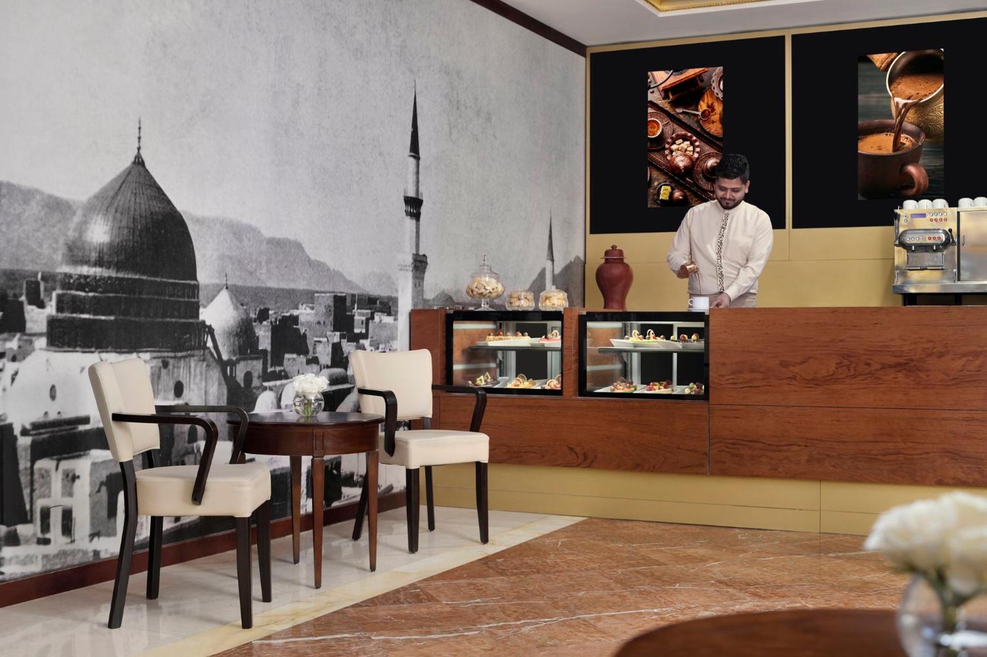 Intercontinental Dar Al Iman Madinah By Ihg Hotel Medina Kültér fotó The photo shows a stylish interior of a café or pastry shop. In the background, there is a large black-and-white mural depicting a cityscape, featuring a prominent dome and minaret, suggesting an architectural style that could be from a Middle Easter