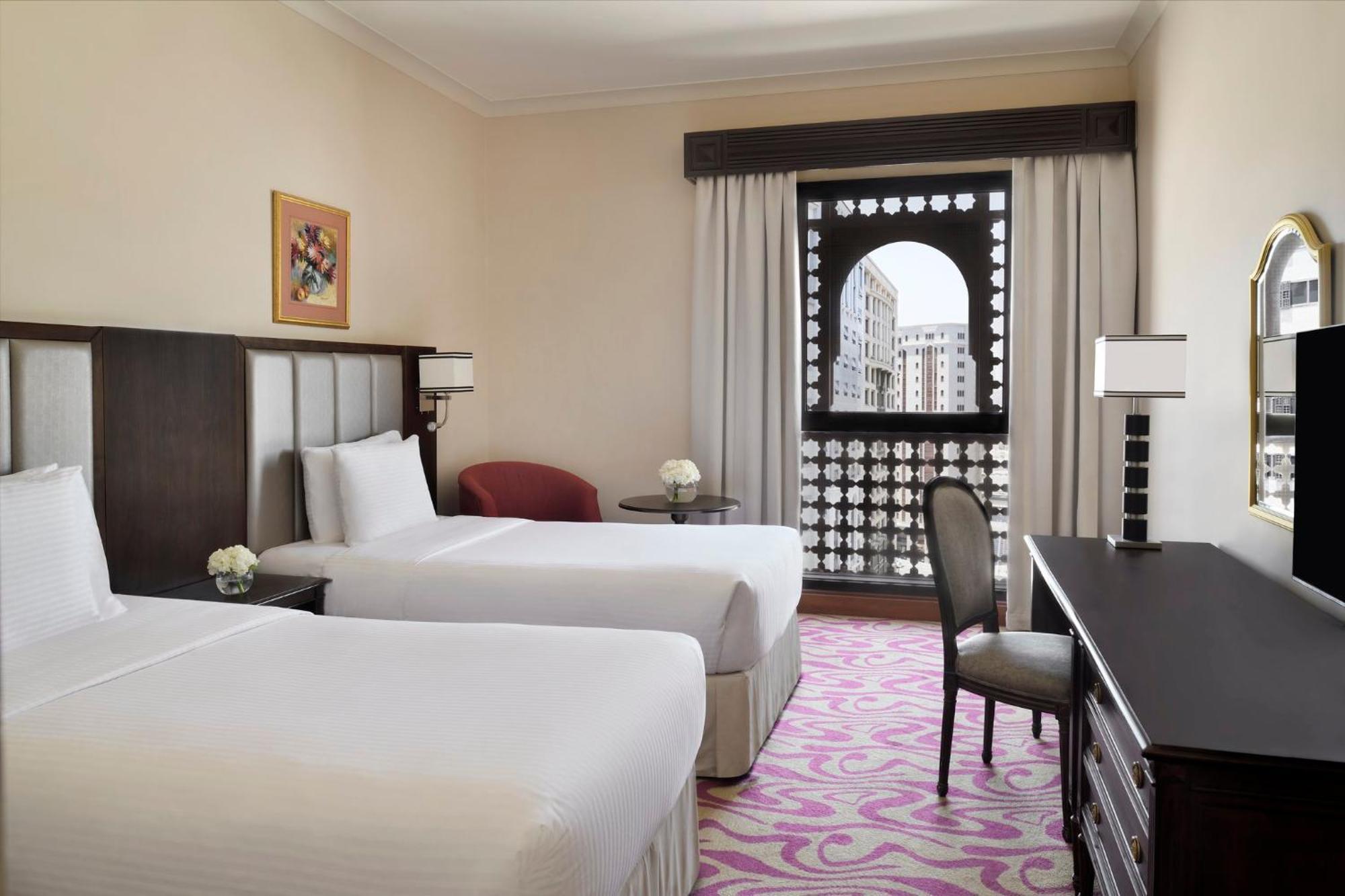 Intercontinental Dar Al Iman Madinah By Ihg Hotel Medina Kültér fotó The photo shows a hotel room featuring two single beds with white bedding. The room has a light, neutral color scheme and is decorated with minimal furnishings. There's a small seating area with a red chair and a round table, as well as a desk with a