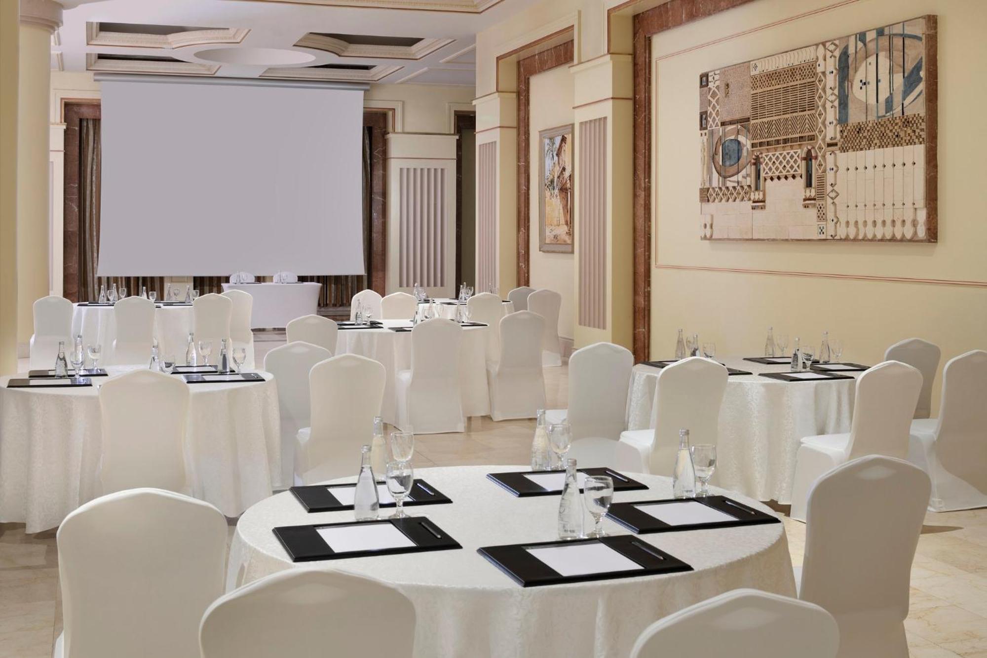 Intercontinental Dar Al Iman Madinah By Ihg Hotel Medina Kültér fotó The photo shows a well-decorated event space or meeting room. It features several round tables covered with white tablecloths and surrounded by white chairs. Each table has black placemats and glass water bottles, suggesting a formal setup for a gath