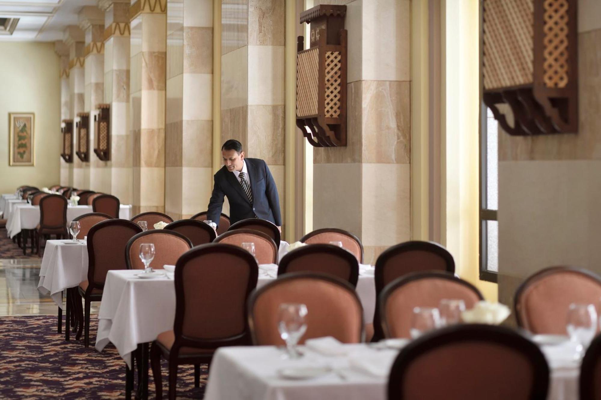 Intercontinental Dar Al Iman Madinah By Ihg Hotel Medina Kültér fotó The photo depicts a dining area in an elegant restaurant. There are several tables set with white tablecloths, each accompanied by chairs. A waiter, dressed in a formal suit, is seen arranging or tending to one of the tables. The restaurant features 