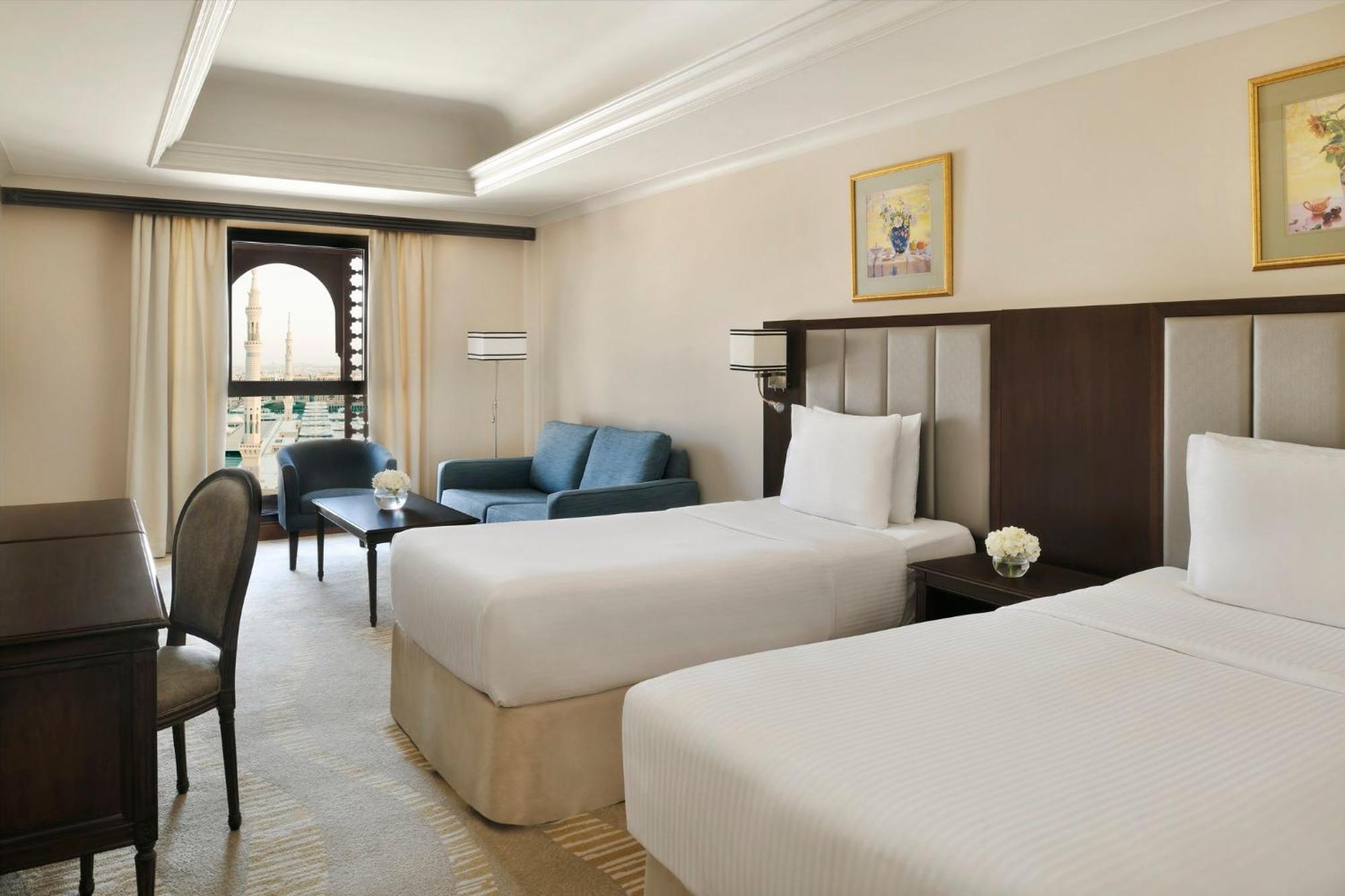 Intercontinental Dar Al Iman Madinah By Ihg Hotel Medina Kültér fotó The photo shows a well-appointed hotel room featuring two twin beds with white bedding. There is a small desk with a chair, a comfortable blue sofa, and a coffee table in the sitting area. The room has a warm, neutral color scheme, accented by two pa