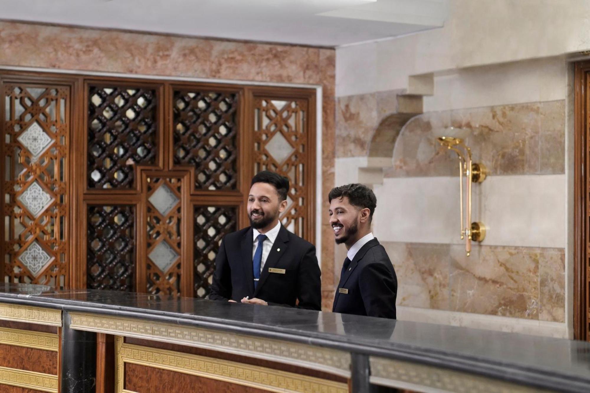 Intercontinental Dar Al Iman Madinah By Ihg Hotel Medina Kültér fotó The photo shows two men standing behind a reception desk in an elegant setting, likely a hotel or similar establishment. They are both dressed in formal attire, wearing suits and ties, and appear to be smiling or engaging in conversation. The backgro