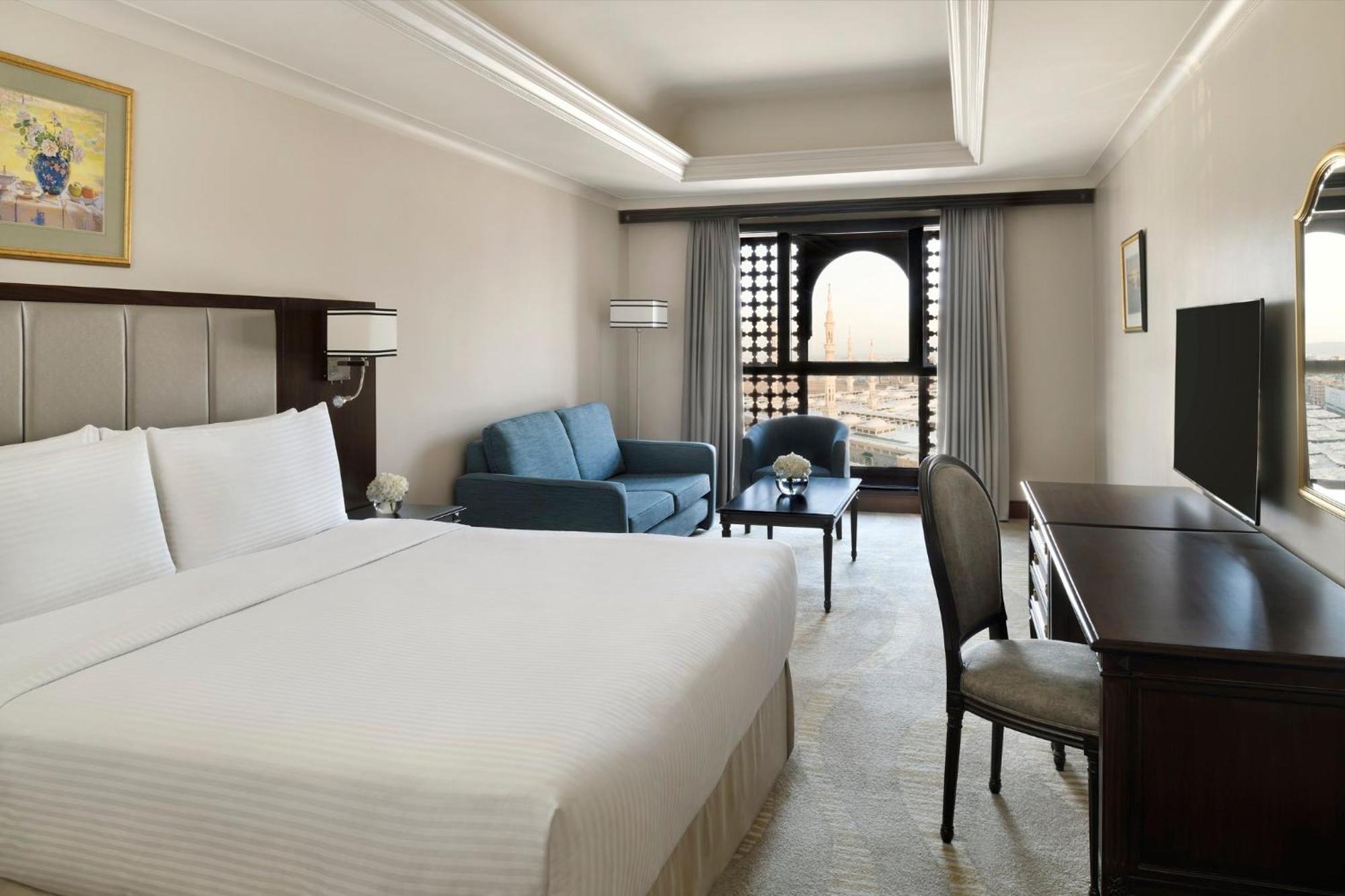Intercontinental Dar Al Iman Madinah By Ihg Hotel Medina Kültér fotó The photo shows a hotel room designed with a modern and comfortable aesthetic. It features a large bed with white linens, a seating area with a blue sofa, and a small table. There is a wooden desk with a chair and a television opposite the bed. Natur