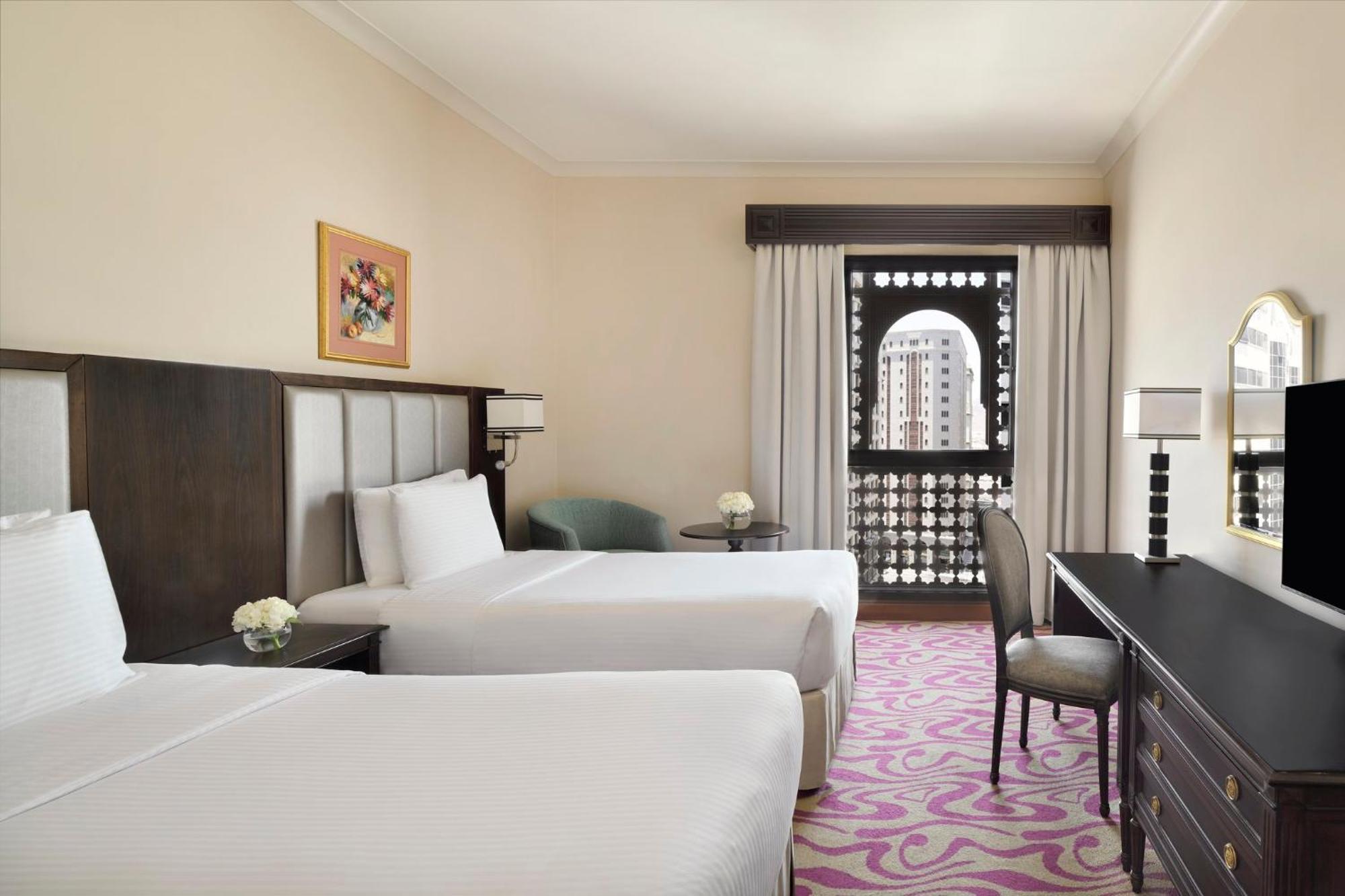 Intercontinental Dar Al Iman Madinah By Ihg Hotel Medina Kültér fotó The photo shows a hotel room featuring two twin beds with white bedding. There is a decorative piece of art on the wall above one of the beds. A small round table with a flower arrangement is placed next to the beds. A desk and chair are situated aga
