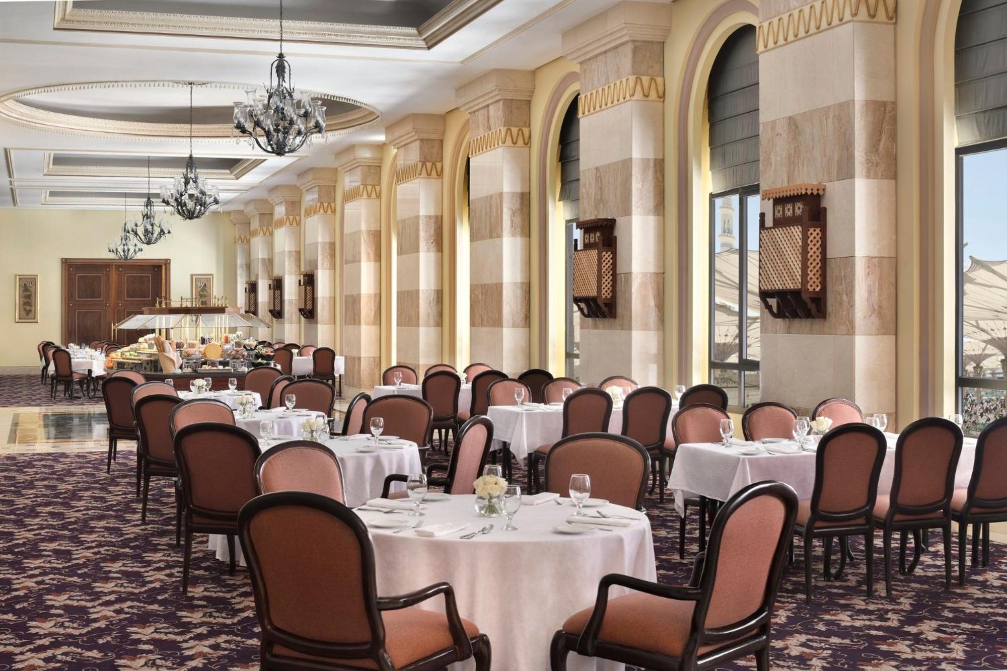 Intercontinental Dar Al Iman Madinah By Ihg Hotel Medina Kültér fotó The photo depicts an elegant dining room setting. It features several round tables covered with white tablecloths, each set with dishes and glassware. The room has a sophisticated ambiance, characterized by high ceilings adorned with chandeliers and 