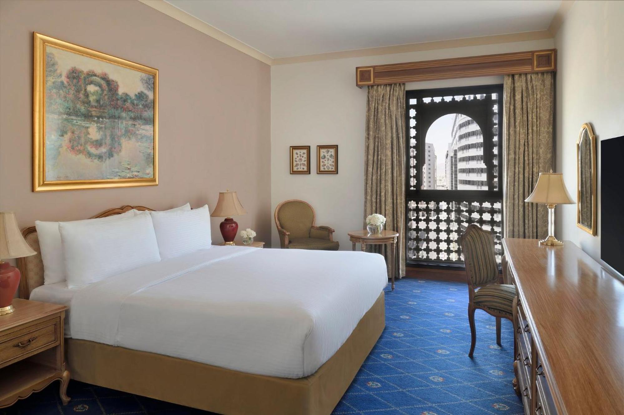 Intercontinental Dar Al Iman Madinah By Ihg Hotel Medina Kültér fotó The photo shows a hotel room that features a large, neatly made bed with white linens. There is a framed painting on the wall above the bed. To the side, a small seating area includes a chair and a side table. Opposite the bed, there is a wooden desk