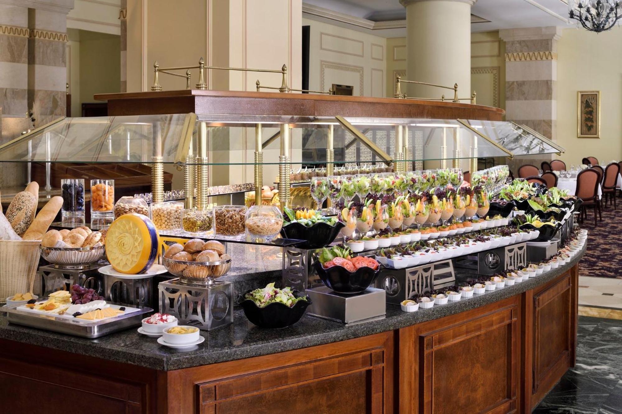 Intercontinental Dar Al Iman Madinah By Ihg Hotel Medina Kültér fotó The photo shows an elegantly arranged buffet with a variety of food items. There is a large, curved wooden display with glass covers, showcasing an assortment of dishes. These include different types of breads and rolls, salads, garnishes, and desser