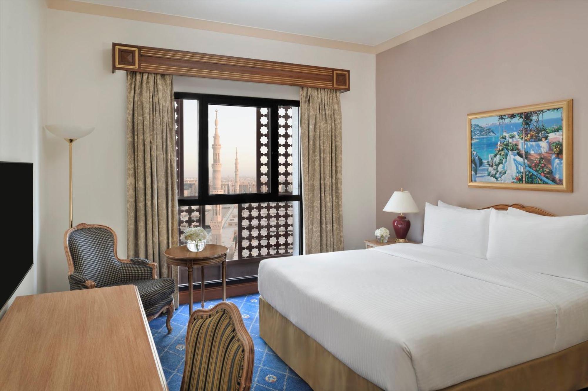 Intercontinental Dar Al Iman Madinah By Ihg Hotel Medina Kültér fotó The photo shows a hotel room with a modern design. There is a large bed with white linens placed against the wall. Next to the bed, there is a small bedside table with a lamp. A comfortable armchair is situated nearby, accompanied by a wooden desk. T
