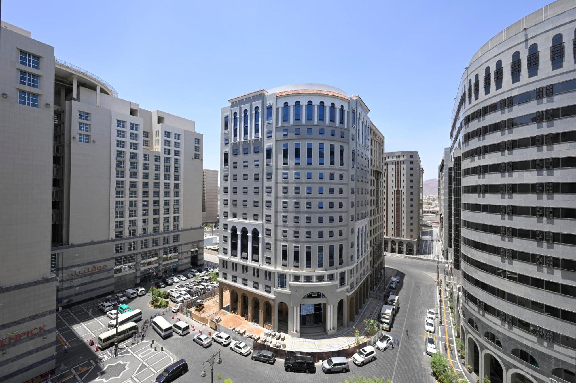 Intercontinental Dar Al Iman Madinah By Ihg Hotel Medina Kültér fotó The photo shows a modern urban scene featuring several buildings, including a prominent rounded building with a glass façade. This building appears to be situated at an intersection, surrounded by other high-rise structures that have distinct archite