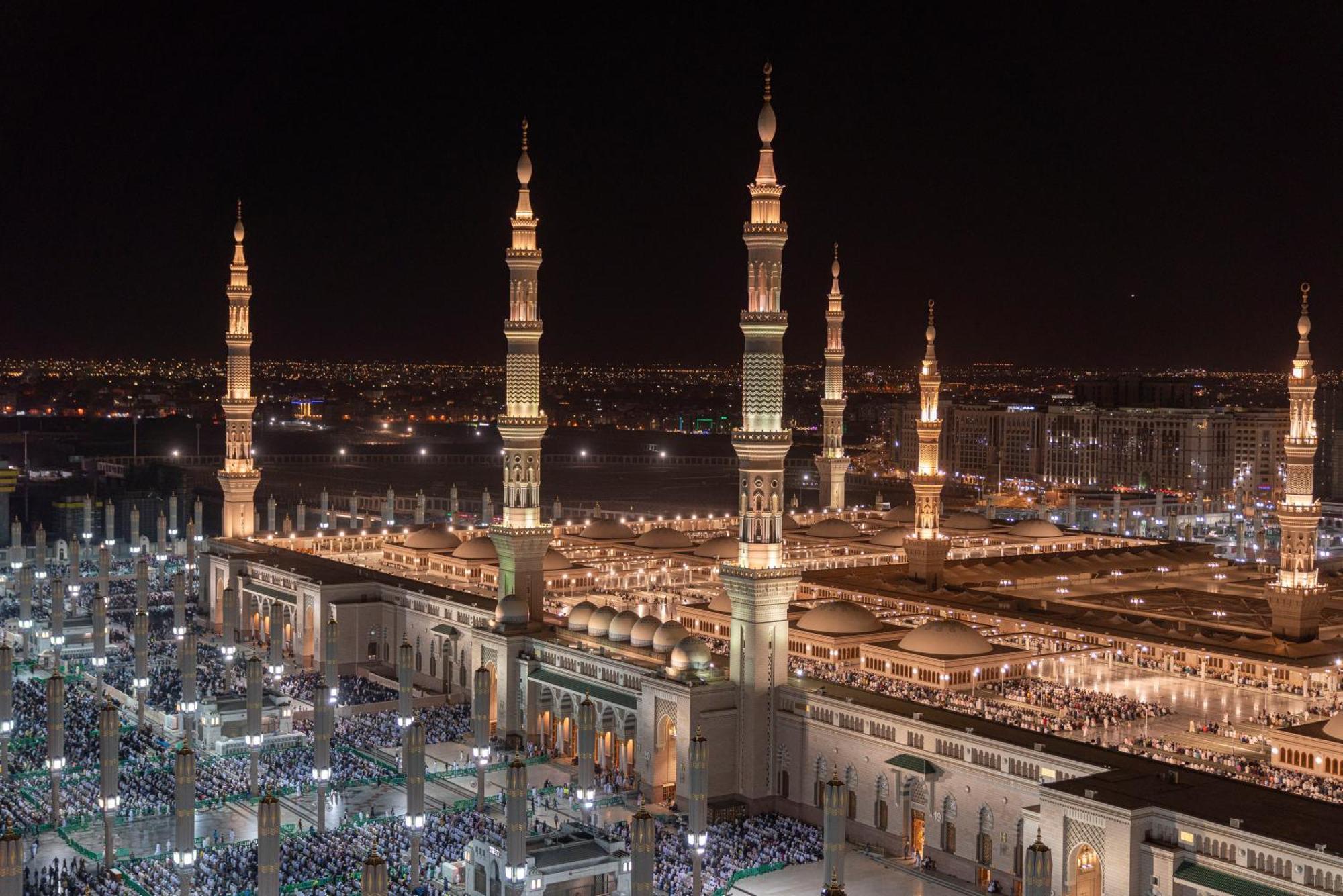 Intercontinental Dar Al Iman Madinah By Ihg Hotel Medina Kültér fotó The photo depicts a stunning nighttime view of a large mosque complex, prominently featuring tall minarets that are illuminated against the dark sky. The surrounding area is bustling with people, likely worshippers, gathered in the courtyard of the m