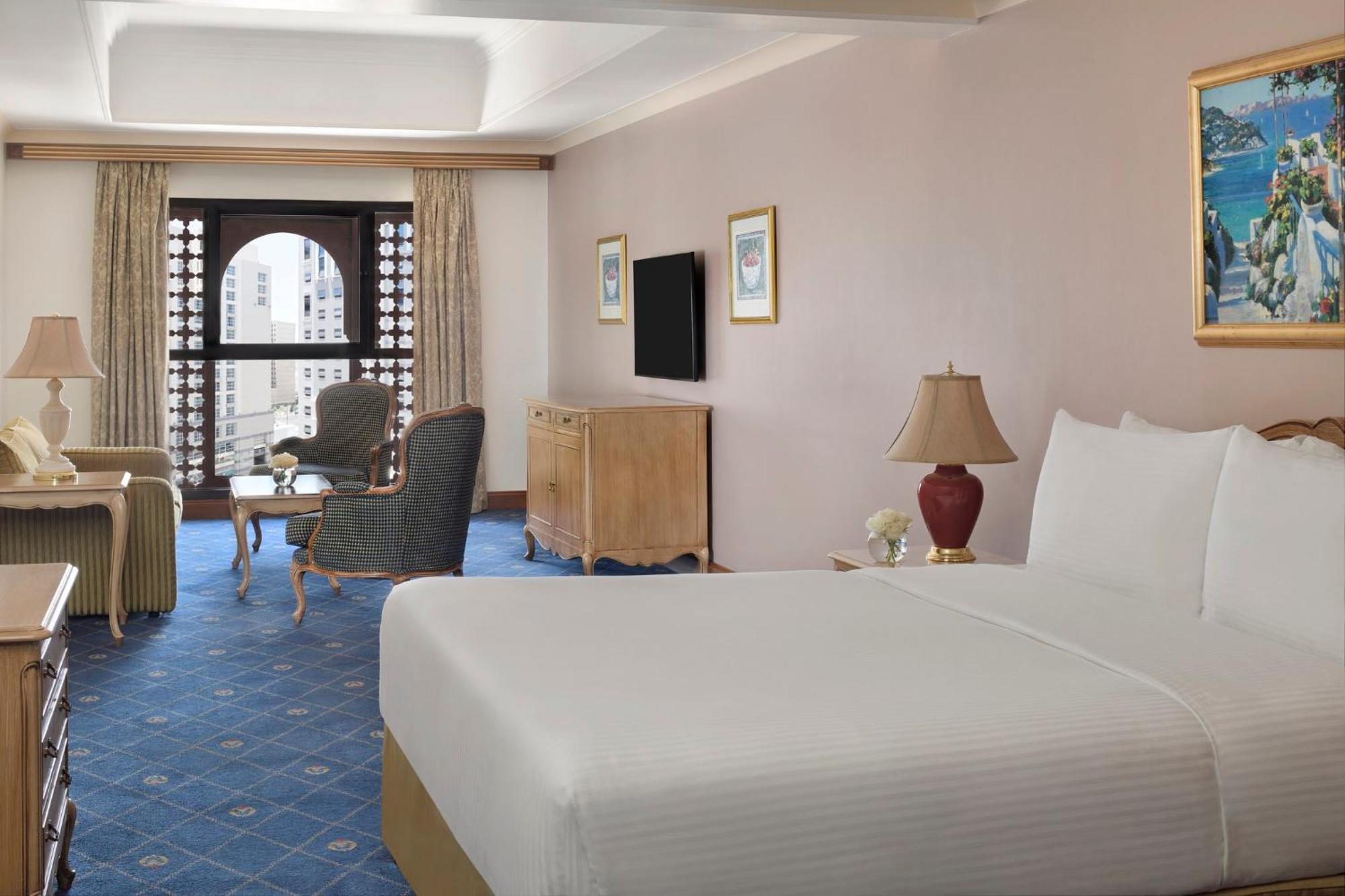 Intercontinental Dar Al Iman Madinah By Ihg Hotel Medina Kültér fotó The photo shows a well-furnished hotel room. It features a large bed with a white duvet, two bedside lamps, and decorative paintings on the walls. There is a comfortable seating area with two chairs and a small table. A wooden dresser is positioned a