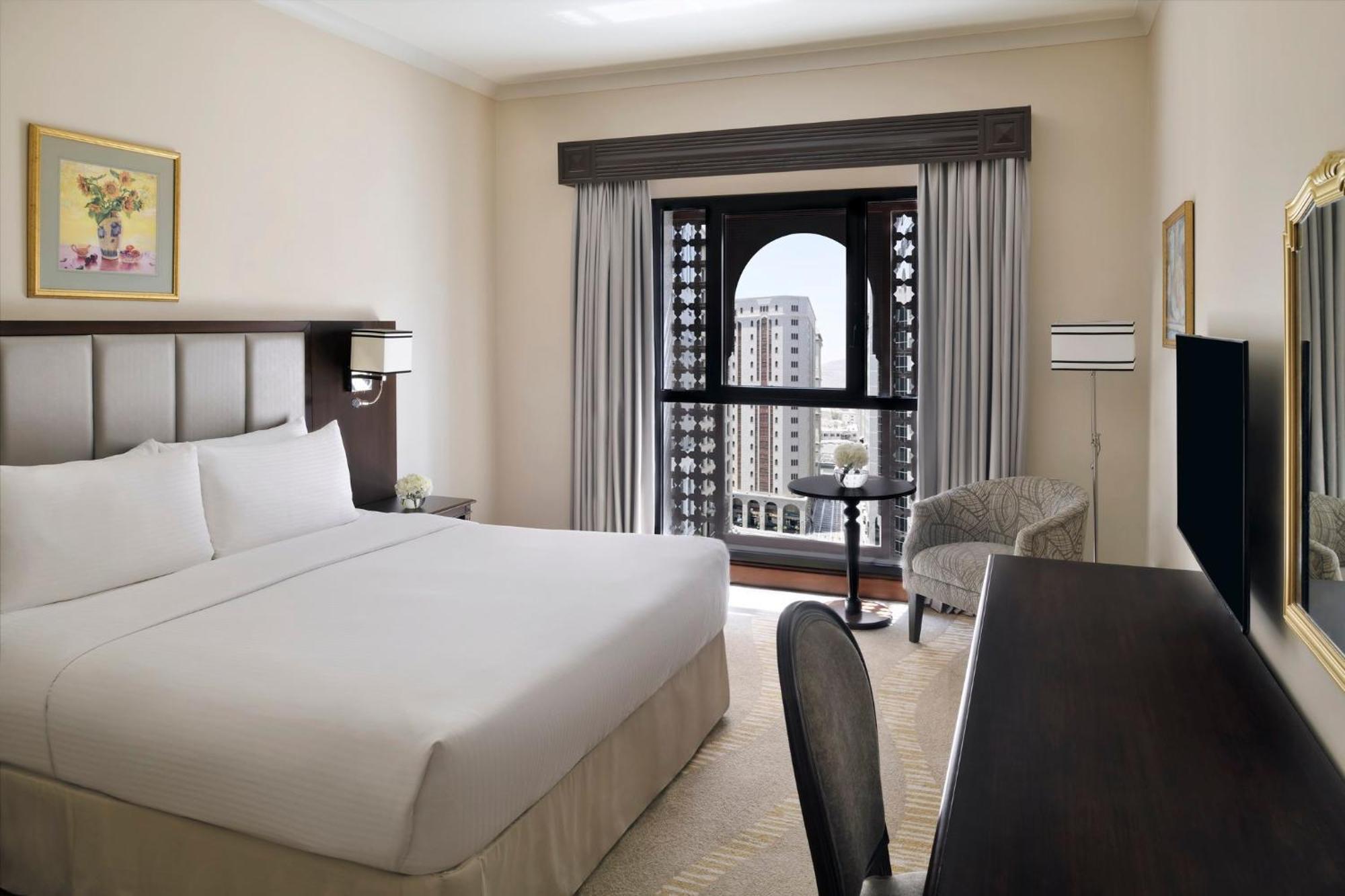 Intercontinental Dar Al Iman Madinah By Ihg Hotel Medina Kültér fotó The photo shows a hotel room with a modern and minimalistic design. It features a large bed with white linens, a side table, and a comfortable chair near a window. The window has a view of a cityscape, and elegant curtains frame the window. There is 