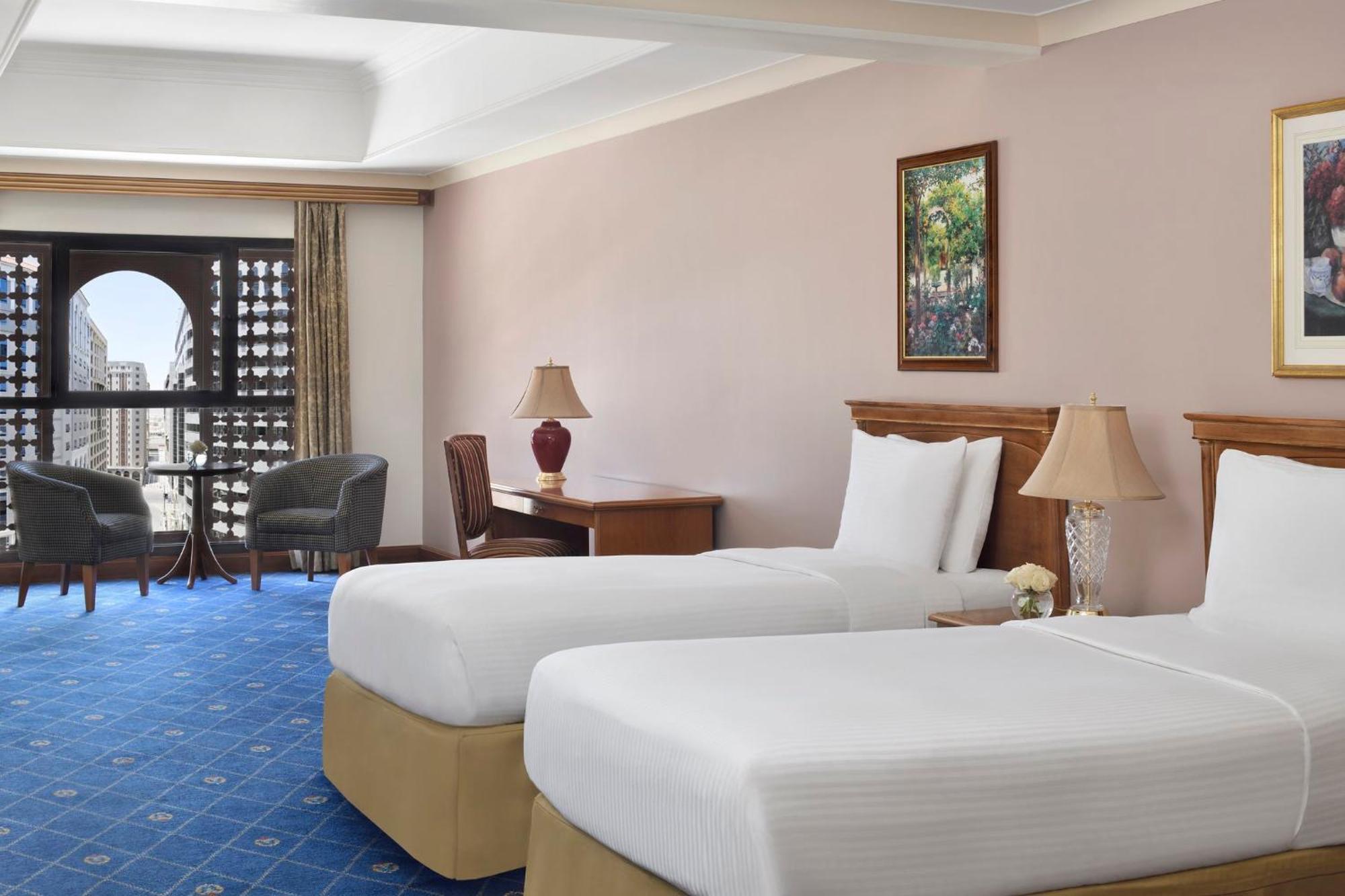 Intercontinental Dar Al Iman Madinah By Ihg Hotel Medina Kültér fotó The photo shows a nicely designed hotel room featuring two twin beds with white linens and beige bases. In the background, there's a wooden desk and a lamp, along with a pair of comfortable armchairs positioned near a large window. The window showcas