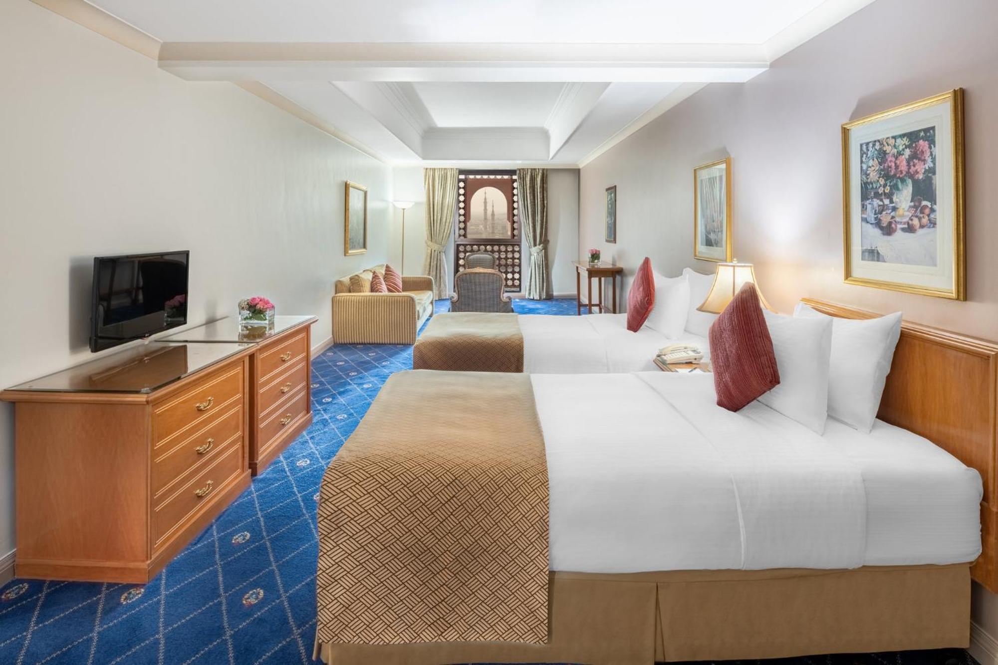 Intercontinental Dar Al Iman Madinah By Ihg Hotel Medina Kültér fotó The photo shows a hotel room featuring two beds with white linens and decorative pillows. There is a wooden dresser with a television on it. The flooring has a blue carpet with a pattern, and the walls are painted in a light color. The room is well-l