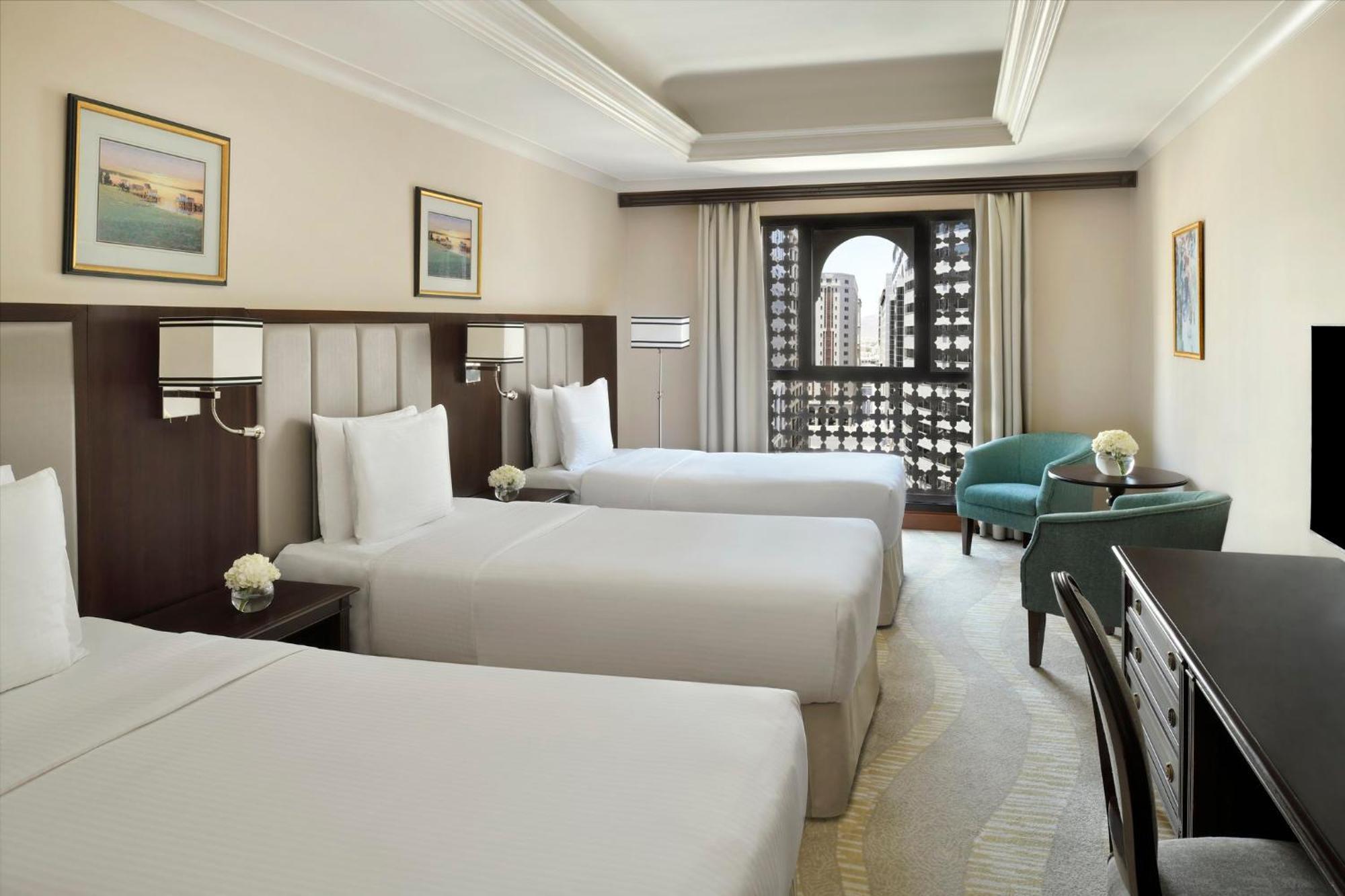 Intercontinental Dar Al Iman Madinah By Ihg Hotel Medina Kültér fotó The photo shows a hotel room with three single beds, dressed in white linens. There are two paintings on the walls, depicting serene landscapes. A large window allows natural light to enter, and it is adorned with elegant curtains. A smaller seating 