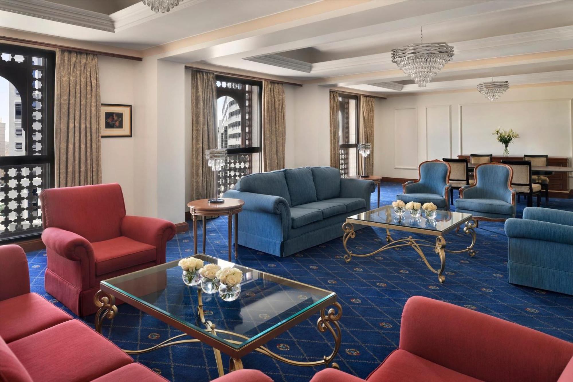 Intercontinental Dar Al Iman Madinah By Ihg Hotel Medina Kültér fotó The photo shows a luxurious living space or lounge area. It features a combination of furniture, including a blue sofa, several red armchairs, and glass-top coffee tables with gold accents. The flooring is carpeted in a deep blue, and the walls are a