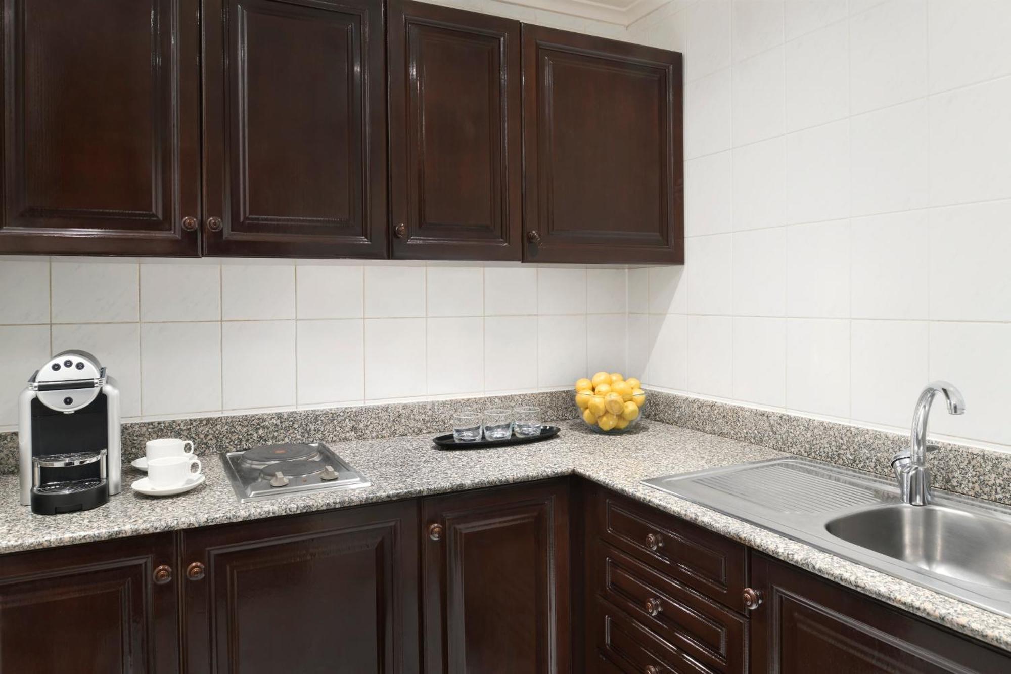 Intercontinental Dar Al Iman Madinah By Ihg Hotel Medina Kültér fotó The photo shows a kitchen with dark wooden cabinetry and a light-colored countertop. There are a few appliances visible, including a coffee machine and a stovetop with burners. To the right, there is a sink, and a bowl of yellow lemons is placed on t