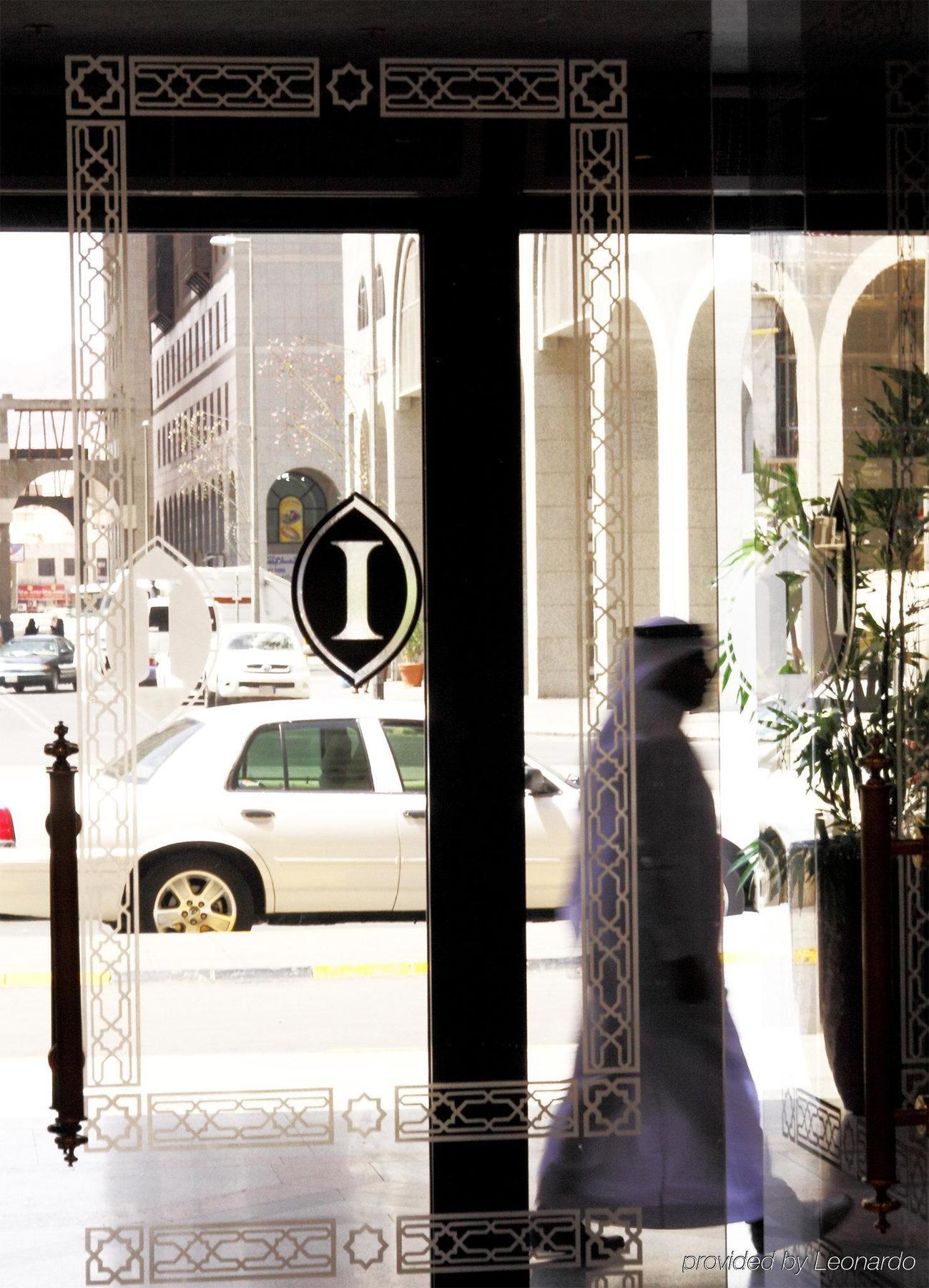 Intercontinental Dar Al Iman Madinah By Ihg Hotel Medina Beltér fotó The photo shows a view through a glass door or window, featuring an elegant design with a prominent "I" symbol displayed. Through the glass, there are some parked vehicles visible, including a white car. In the foreground, a shadowy figure of a perso