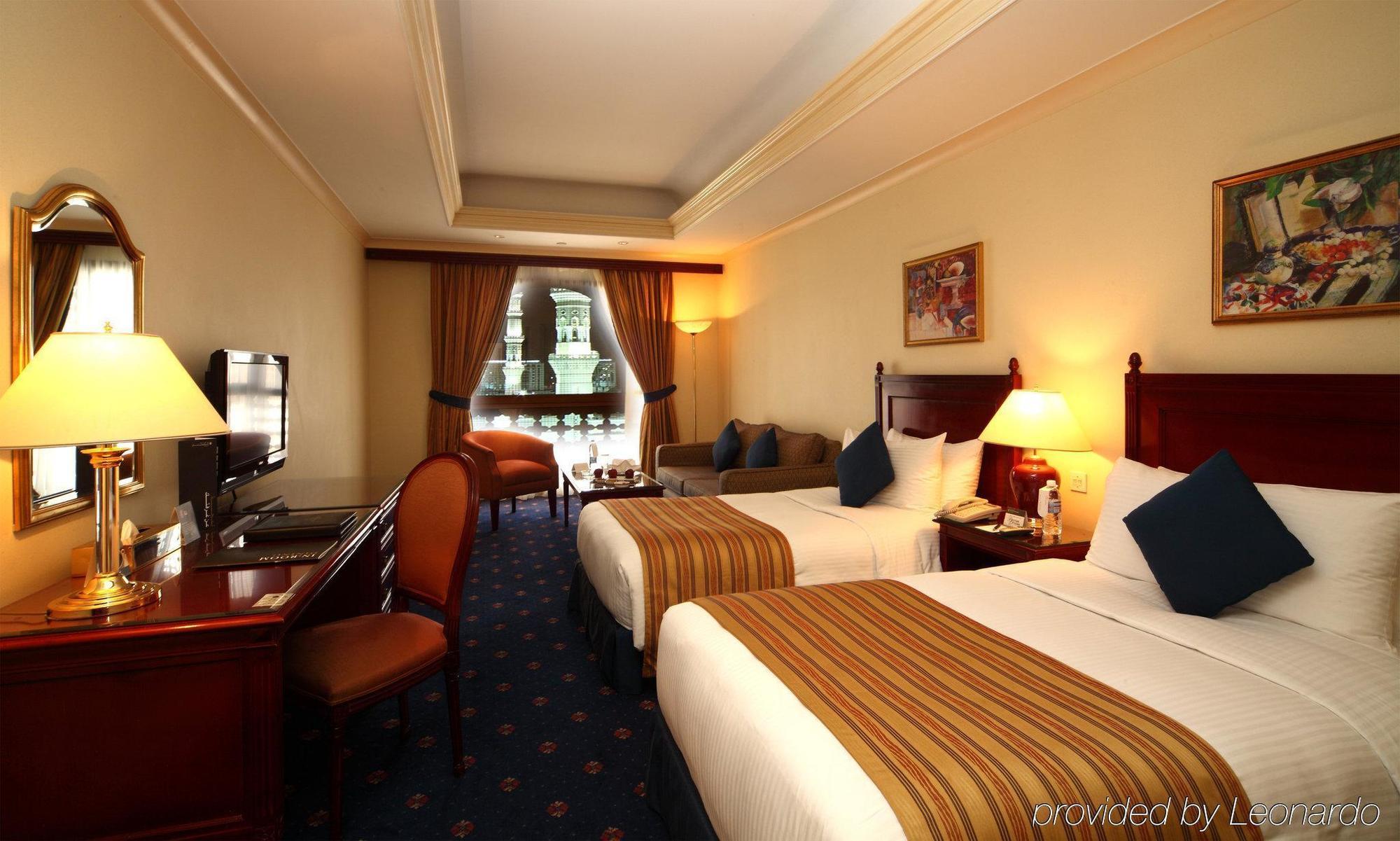 Intercontinental Dar Al Iman Madinah By Ihg Hotel Medina Szoba fotó The photo shows a hotel room featuring two beds with white linens and brown striped throws. There is a wooden desk and chair, alongside a television. The walls are painted in a warm tone, and there are decorative paintings. Large windows with curtain