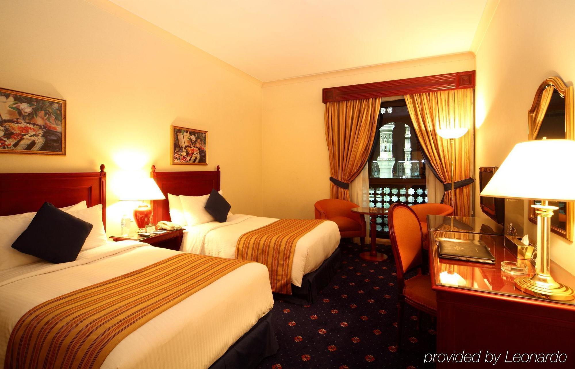 Intercontinental Dar Al Iman Madinah By Ihg Hotel Medina Szoba fotó The photo shows a hotel room featuring two twin beds with white linens and brown striped bedspreads. There are decorative pillows on the beds, and the room has warm-colored walls. To the side, there's a small sitting area with a round table and a cha