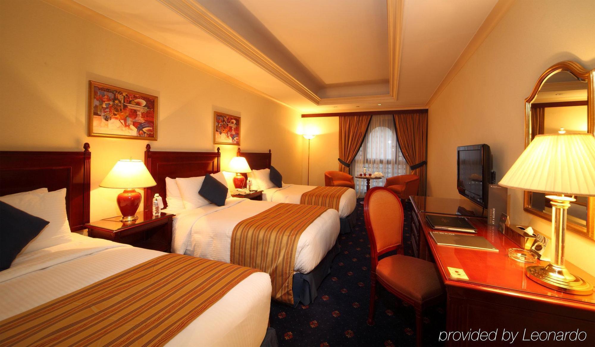 Intercontinental Dar Al Iman Madinah By Ihg Hotel Medina Szoba fotó The photo shows a hotel room with a warm and inviting atmosphere. There are two beds with white and gold bedding, complemented by decorative pillows. Each bed has a bedside lamp. In the background, there's a small seating area with a couple of chairs