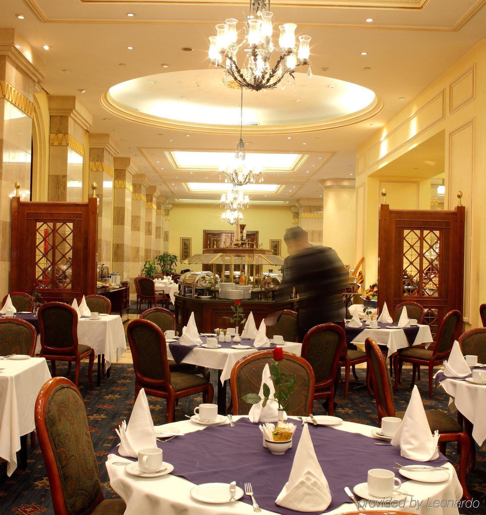 Intercontinental Dar Al Iman Madinah By Ihg Hotel Medina Étterem fotó The photo depicts an elegant restaurant interior. It features round tables covered with white tablecloths, each set with plates, cutlery, and folded napkins arranged into triangles. The space has a warm, inviting atmosphere with a mix of decorative e