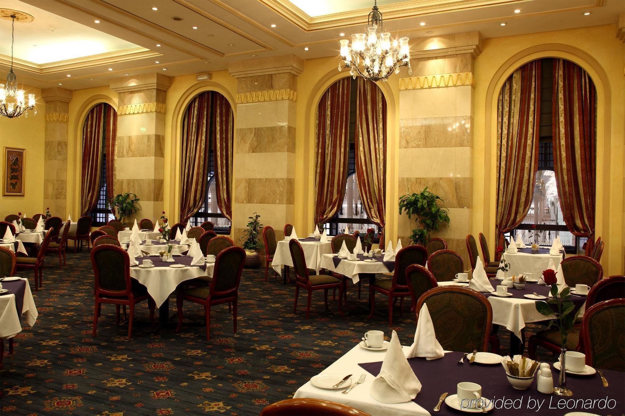 Intercontinental Dar Al Iman Madinah By Ihg Hotel Medina Étterem fotó The photo depicts an elegant restaurant interior. The space features ornate decor with tall windows draped in rich, patterned curtains. Tables are neatly set with white tablecloths and napkins, and there are plush chairs arranged around them. The flo