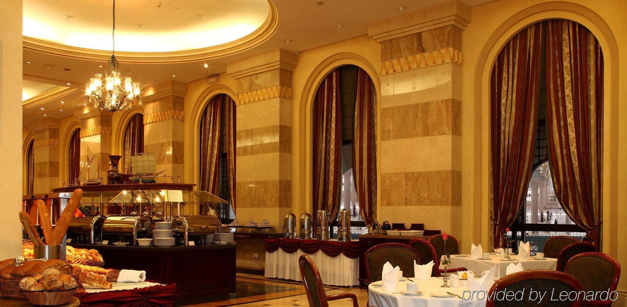 Intercontinental Dar Al Iman Madinah By Ihg Hotel Medina Étterem fotó The photo shows an elegant dining restaurant interior. It features stylish arches and walls with a blend of marble and decorative elements. There are large windows draped with rich curtains, contributing to the luxurious atmosphere. The dining area i