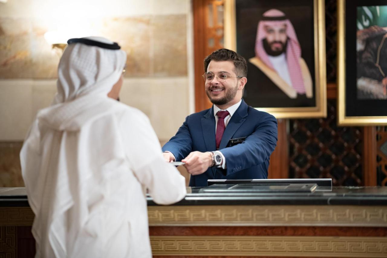 Intercontinental Dar Al Iman Madinah By Ihg Hotel Medina Kültér fotó The photo shows a hotel reception area. A male receptionist, dressed in a navy suit with a tie, is smiling and handing an item to a guest who is wearing traditional white attire, possibly a kandura or thobe. The background features elegantly decorate