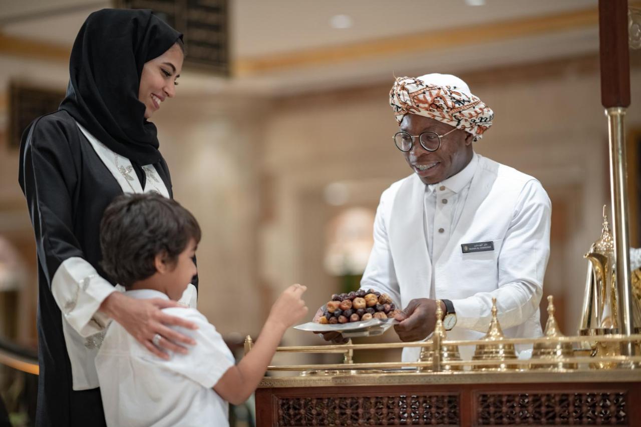 Intercontinental Dar Al Iman Madinah By Ihg Hotel Medina Kültér fotó The photo depicts a warm interaction between three people. A woman, dressed in a traditional black attire and a headscarf, stands beside a young boy who is reaching out towards a plate of dates held by a smiling man in a white traditional outfit with